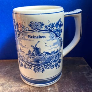 May include: White ceramic mug with a blue and white Dutch windmill design and the word "Heineken". The mug has a handle and a blue rim. The design includes floral patterns and a landscape scene.