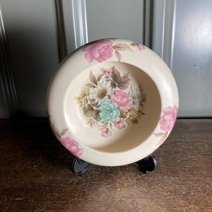 Könnte beinhalten: Eine cremefarbene, runde dekorative Schale mit floralem Design. Die Schale zeigt rosa, blaue und weiße Blumen. Die Schale wird auf einem schwarzen Ständer präsentiert.