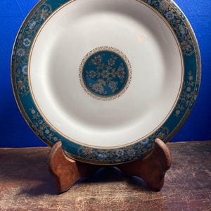 Può includere: Un piatto in ceramica bianca con bordo e motivo centrale floreale verde acqua e oro. Il piatto è esposto su un supporto in legno su sfondo blu. Il bordo del piatto presenta un motivo floreale ripetuto con dettagli dorati.