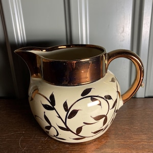 May include: Cream-colored ceramic pitcher with a bronze-colored rim, handle, and spout. The pitcher features a decorative pattern of dark brown leaves and vines. The handle is curved, and the spout is designed for easy pouring.