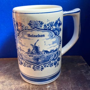 May include: White ceramic mug with a blue and white Dutch windmill design and the word "Heineken". The mug has a handle and a blue border at the top and bottom. The mug is on a wooden surface.