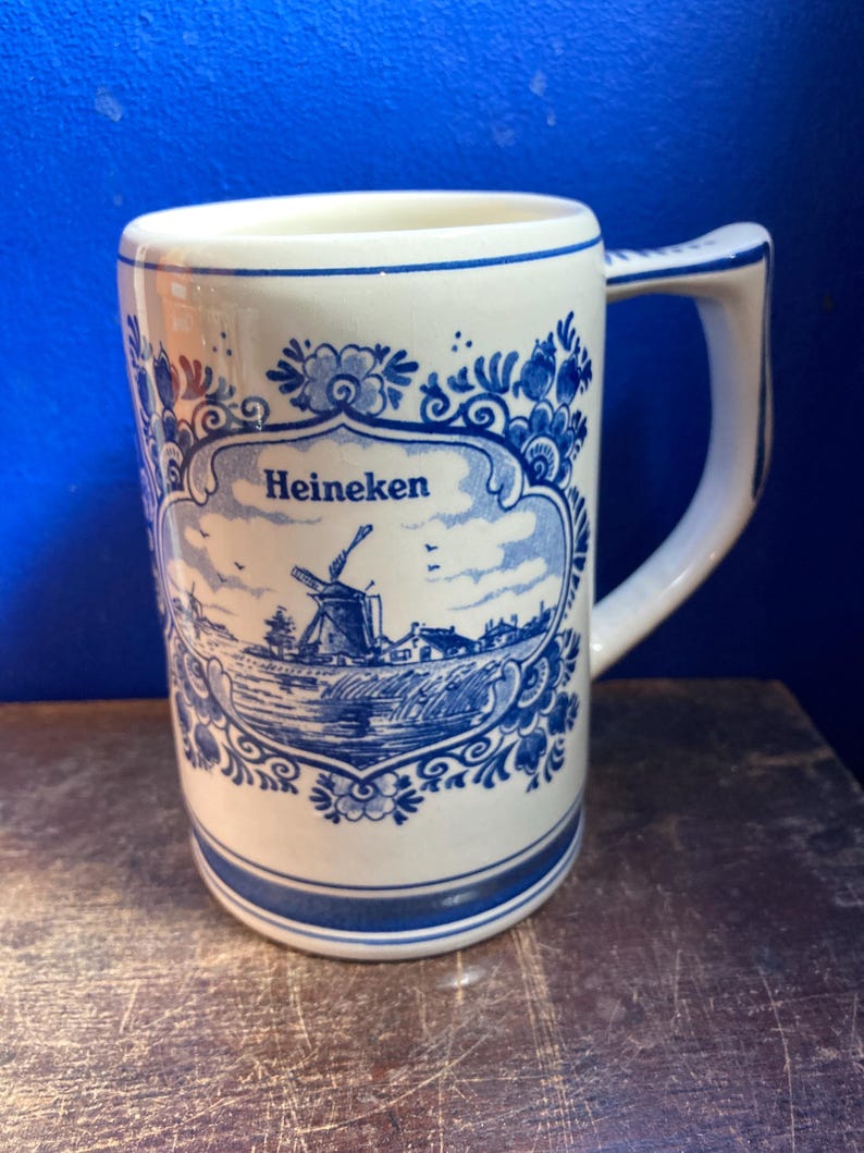 May include: White ceramic mug with a blue and white Dutch windmill design and the word "Heineken". The mug has a handle and a blue border at the top and bottom. The mug is on a wooden surface.