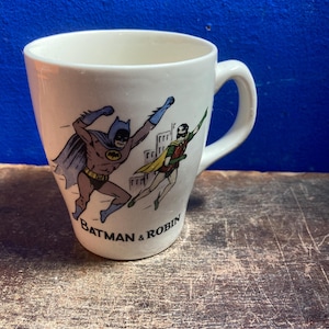 May include: White ceramic mug featuring Batman and Robin in action poses. The mug has a curved shape and a handle. The words "Batman & Robin" are printed below the illustrations. The background is a distressed wooden surface.