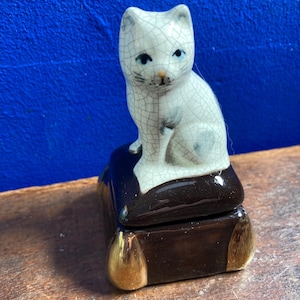 Puede incluir: Figura de gato de cerámica con acabado craquelado, sobre una caja rectangular marrón oscuro con detalles dorados. El gato tiene ojos azules y está sentado erguido. La caja parece una caja de baratijas o joyero.