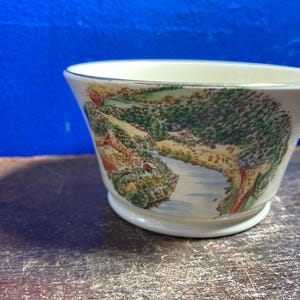 May include: A small, off-white ceramic bowl with a landscape scene. The bowl features a detailed painting of a river, trees, and a small building. The rim is trimmed with a gold band. The bowl sits on a wooden surface.