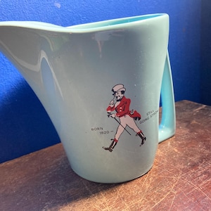 May include: A light blue ceramic pitcher with a handle. The pitcher features a graphic of a man in a red coat and the text "Born 1820 - Still Going Strong."