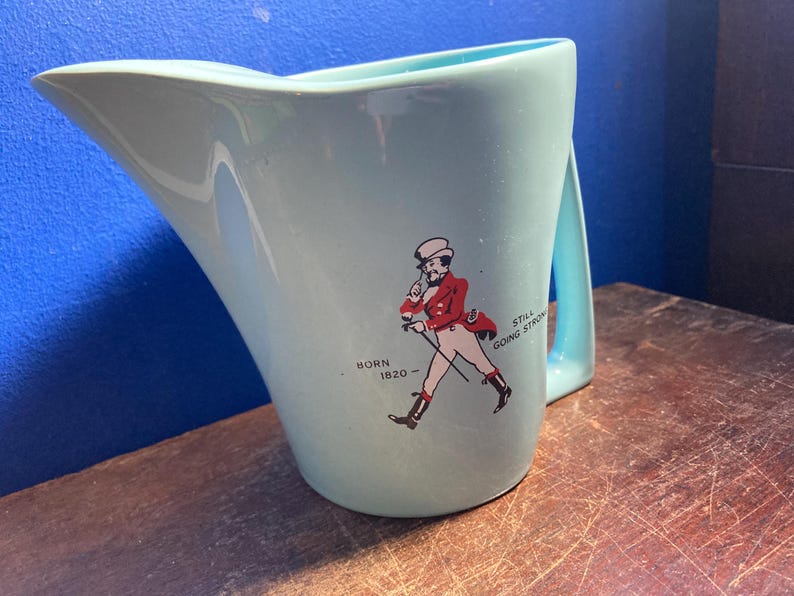 May include: A light blue ceramic pitcher with a handle. The pitcher features a graphic of a man in a red coat and the text "Born 1820 - Still Going Strong."