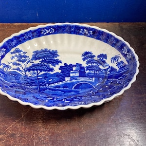 May include: An oval ceramic serving dish featuring a blue and white transferware pattern. The design depicts a landscape with a bridge, buildings, and trees. The rim has a scalloped edge with a floral border, and the interior surface is white.