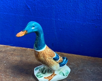 Op de afbeelding: Een keramieken eendenbeeldje met een blauwe kop en nek, bruin lichaam en oranje snavel. De eend staat op een witte basis met groene accenten. Het beeldje is een decoratief item.