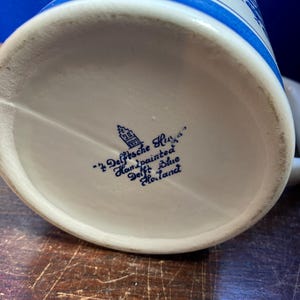May include: A white ceramic beer stein with blue floral patterns and accents. The bottom of the stein features blue text that reads "'t Delfsche Huys Handpainted Delft Blue Holland".