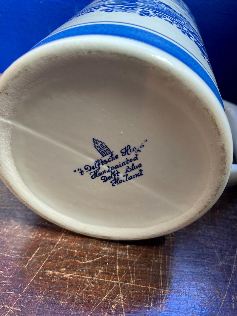 May include: A white ceramic beer stein with blue floral patterns and accents. The bottom of the stein features blue text that reads "'t Delfsche Huys Handpainted Delft Blue Holland".