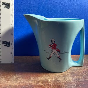 May include: A light blue ceramic pitcher with a curved handle and spout. The pitcher features a graphic of a man in a red coat and the text "BORN 1820 - STILL GOING STRONG". A ruler with black cat graphics is to the left of the pitcher.