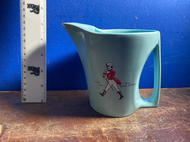 May include: A light blue ceramic pitcher with a curved handle and spout. The pitcher features a graphic of a man in a red coat and the text "BORN 1820 - STILL GOING STRONG". A ruler with black cat graphics is to the left of the pitcher.