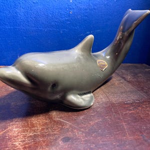 Puede incluir: Una figura de delfín de cerámica gris brillante en posición de nado. El delfín presenta una superficie lisa y reflectante, además de una pequeña pegatina circular de tono dorado en el costado. La escultura muestra al animal con la cola curvada.