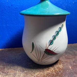 以下が含まれることがあります： ターコイズの蓋が付いた白いセラミックジャー。ジャーは、緑、赤、茶色の花の模様で装飾されています。ジャーは、青い背景に対して木製の表面に置かれています。