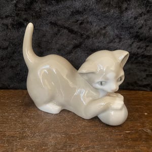 Può includere: Una statuetta in ceramica di un gatto giocoso di colore grigio chiaro, con una palla bianca. Il gatto è in posa giocosa, con le zampe sulla palla. Il gatto ha gli occhi verdi e una coda ricurva.