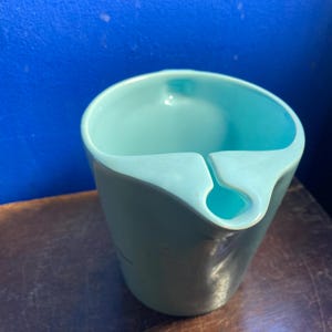 May include: A light teal-colored ceramic cup with a unique design. The cup has a curved rim with a notch and a small hole. The cup is sitting on a brown surface with a blue background.