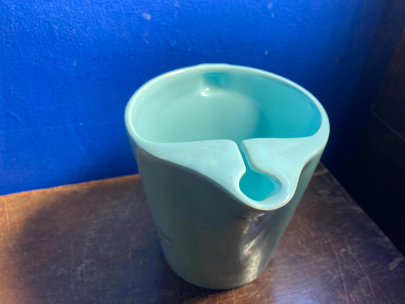 May include: A light teal-colored ceramic cup with a unique design. The cup has a curved rim with a notch and a small hole. The cup is sitting on a brown surface with a blue background.