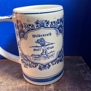 May include: A white ceramic mug with a blue floral design and the words "Wildenrath Golf Club." The mug has a handle and a blue stripe around the top and bottom. The design features a golfer in action.