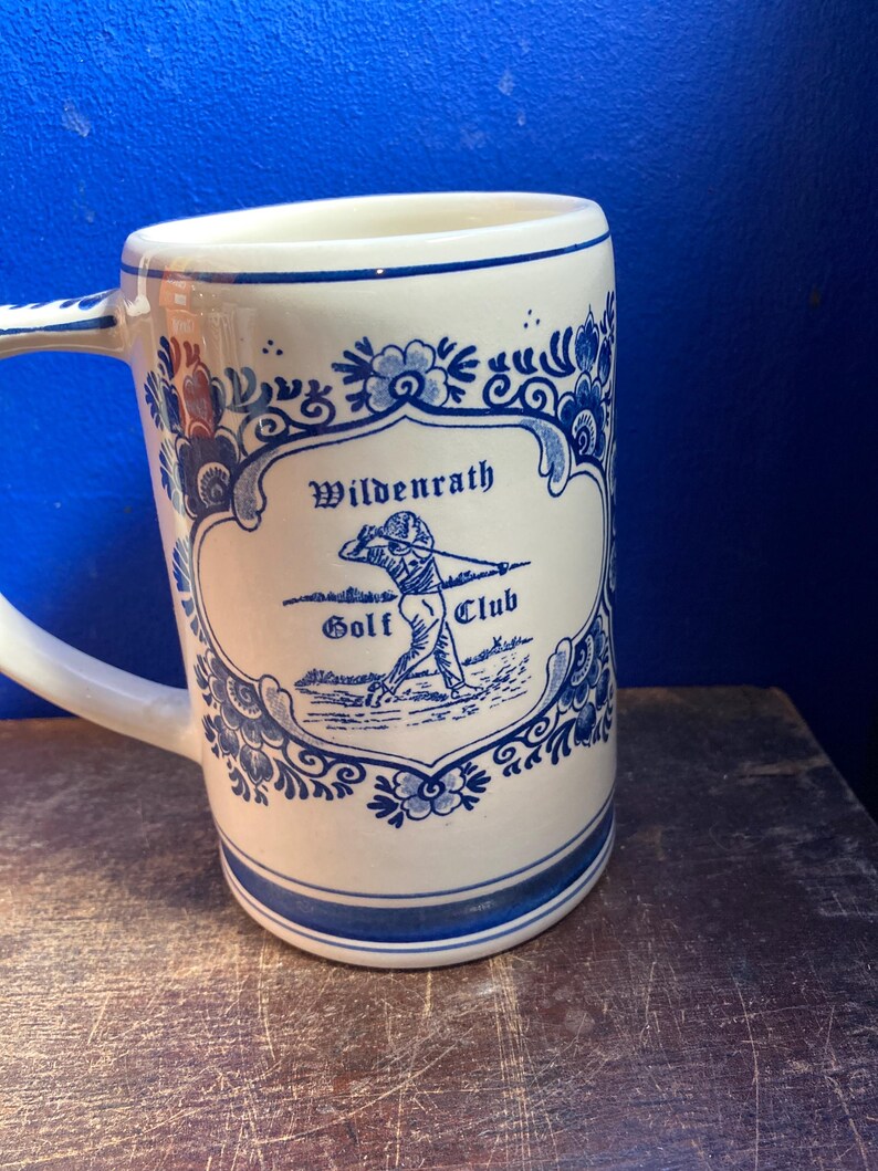 May include: A white ceramic mug with a blue floral design and the words "Wildenrath Golf Club." The mug has a handle and a blue stripe around the top and bottom. The design features a golfer in action.