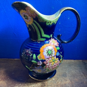 Peut inclure: Un pichet en céramique bleu foncé orné d'un motif floral vibrant avec des fleurs orange, roses et jaunes. Le pichet possède une anse incurvée, un large bec verseur et un bord ainsi qu'une base dorés. La surface présente des feuillages verts.