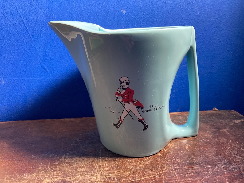May include: A light blue ceramic pitcher with a curved handle and spout. The pitcher features a graphic of a man in a red coat and the text "BORN 1820 - STILL GOING STRONG".