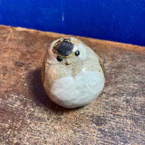 Puede incluir: Una figura de pájaro de cerámica con cuerpo redondeado. El pájaro tiene vientre blanco, lados moteados de color canela y cabeza marrón oscuro. Tiene ojos negros pequeños y un pico ligeramente levantado. La figura está sobre una superficie de madera.