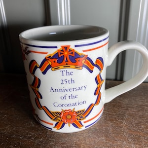 May include: White ceramic mug with a handle, featuring a design commemorating the 25th Anniversary of the Coronation. The design includes a crown, ribbons, and floral elements in orange, blue, and red. The mug has blue and red stripes near the rim.