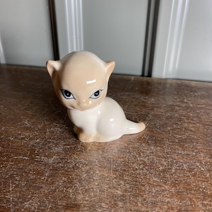 May include: A ceramic figurine of a stylized cat with a cream-colored head and white body. The cat has black eyes and small, pointed ears. The figurine is sitting on a wooden surface.