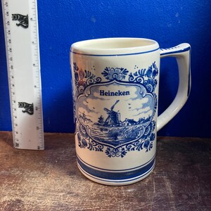 May include: A white ceramic mug with a blue and white windmill design and the word "Heineken". The mug has a handle and a blue border at the top and bottom. The mug is approximately 5 inches tall.