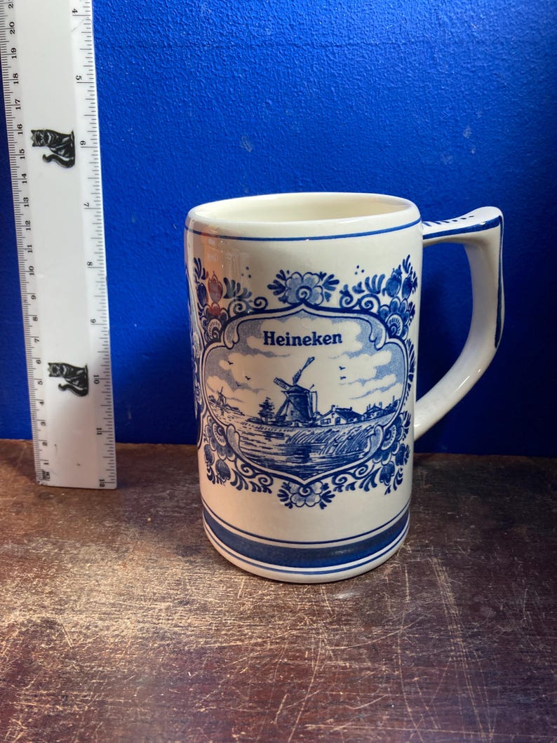 May include: A white ceramic mug with a blue and white windmill design and the word "Heineken". The mug has a handle and a blue border at the top and bottom. The mug is approximately 5 inches tall.