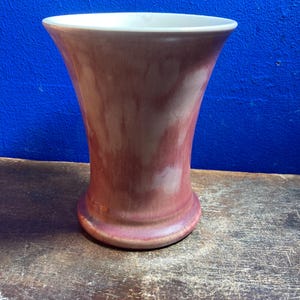 May include: A maroon ceramic vase with a flared top and a rounded base. The vase has a smooth, glossy finish and a white interior. The vase is displayed on a weathered wooden surface.