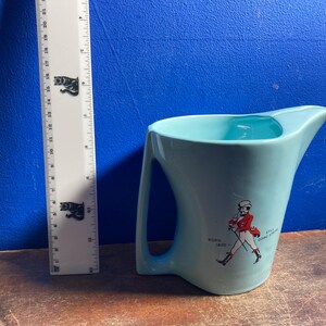 May include: A light blue ceramic pitcher with a handle and a spout. The pitcher has a graphic of a man in a red coat and the text "Born 1820". A ruler is next to the pitcher.