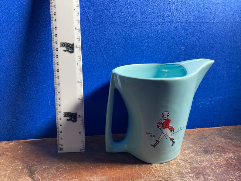 May include: A light blue ceramic pitcher with a handle and a spout. The pitcher has a graphic of a man in a red coat and the text "Born 1820". A ruler is next to the pitcher.