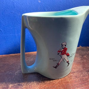 May include: A light blue ceramic pitcher with a curved handle and a spout. The pitcher features a graphic of a man in a red coat and the text "BORN 1820 - STILL GOING STRONG". The interior is a lighter blue.