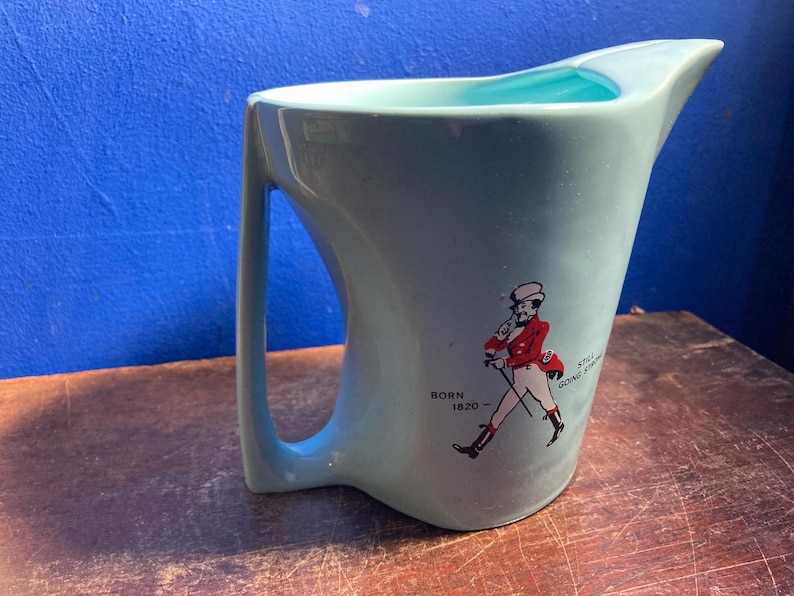 May include: A light blue ceramic pitcher with a curved handle and a spout. The pitcher features a graphic of a man in a red coat and the text "BORN 1820 - STILL GOING STRONG". The interior is a lighter blue.