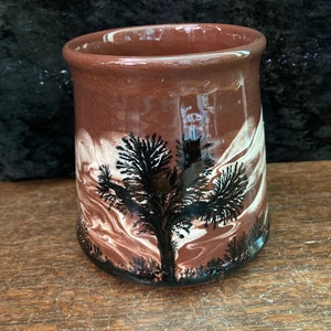 Könnte beinhalten: Eine braune Keramiktasse mit einem weißen und braunen Marmormuster und einer schwarzen Silhouette eines Baumes.