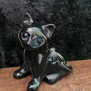 Może przedstawiać: Czarna ceramiczna figurka kota z błyszczącym wykończeniem. Kot siedzi z nogami schowanymi pod sobą i ogonem owiniętym wokół ciała.