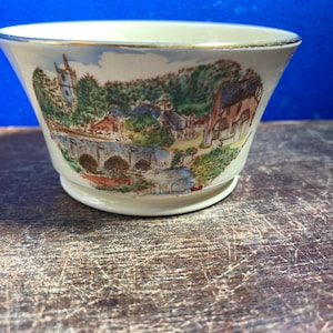 May include: Cream-colored ceramic bowl with a detailed landscape scene featuring a bridge, buildings, and trees. The rim is trimmed with gold. The bowl has a flared shape and sits on a small base.