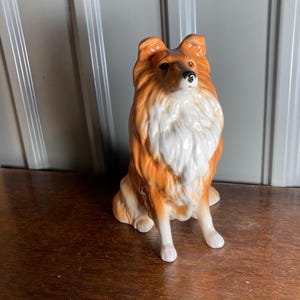 May include: A ceramic Collie dog figurine. The dog has a brown and white coat, with a fluffy mane and tail. The figurine is sitting upright, with its front paws extended. The dog's eyes are dark, and it has a focused expression. The figurine is a decorative item.