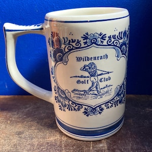 May include: A white ceramic mug with a blue floral design and a handle. The mug features the text "Wildenrath Golf Club" and an illustration of a golfer. The mug has a blue stripe at the base and rim.