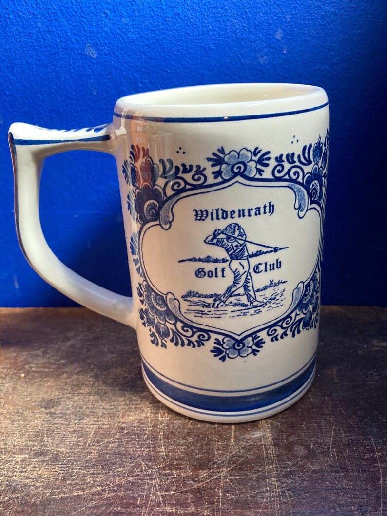 May include: A white ceramic mug with a blue floral design and a handle. The mug features the text "Wildenrath Golf Club" and an illustration of a golfer. The mug has a blue stripe at the base and rim.