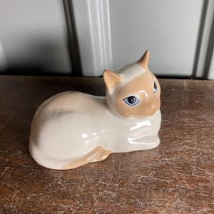 May include: A ceramic figurine of a cat in a resting pose. The cat has a cream-colored body with tan markings on its head, ears, and tail. It has blue eyes and is sitting on a wooden surface.