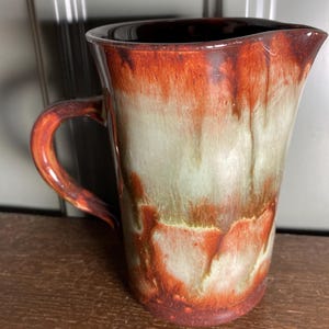 May include: A handmade ceramic jug with a distinctive glaze. The jug has a dark brown rim and handle, blending into red, green, and cream hues. The design has a dripped effect, enhancing its artistic charm. A decorative piece for the home.