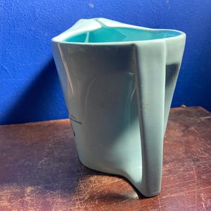 May include: A light blue ceramic pitcher with a unique angular handle and a turquoise interior. The pitcher has a distinctive shape and a small graphic on the side. The pitcher is likely used for serving beverages.