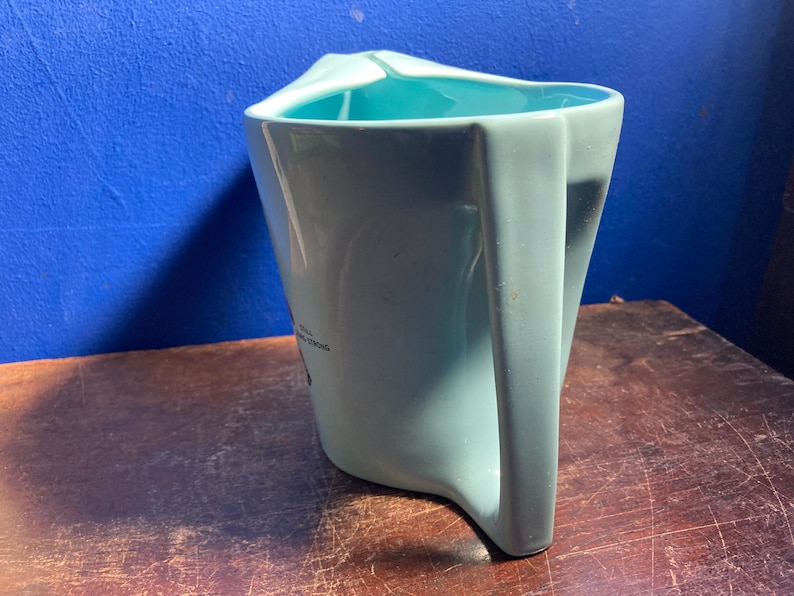 May include: A light blue ceramic pitcher with a unique angular handle and a turquoise interior. The pitcher has a distinctive shape and a small graphic on the side. The pitcher is likely used for serving beverages.