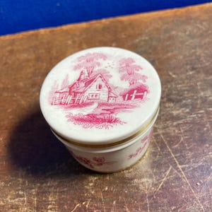 May include: A small, round, white ceramic jar with a lid featuring a red and white pastoral scene. The jar has a gold band around the middle. The lid depicts a house, trees, and a body of water.