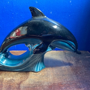 Pode incluir: Uma estatueta de golfinho em cerâmica brilhante com a parte superior do corpo preta e uma secção inferior em azul-petróleo que representa ondas estilizadas. O golfinho é representado numa pose de salto, com a barbatana dorsal virada para cima.