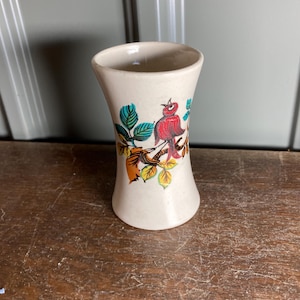 Może przedstawiać: Mały, kremowy ceramiczny wazon z rozszerzaną górą i wąską podstawą. Wazon jest ozdobiony kolorową ilustracją czerwonego ptaka siedzącego na gałęzi z zielonymi, pomarańczowymi i żółtymi liśćmi. Wazon stoi na drewnianej powierzchni.