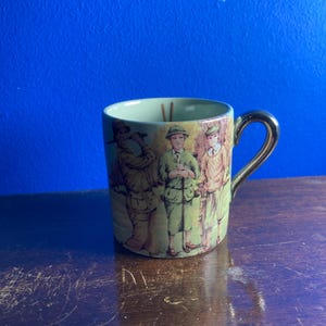 Op de afbeelding: Een vintage keramische mok met een gouden handvat en rand. De mok heeft een geschilderde scène van drie figuren in groene en bruine kleding. De binnenkant van de mok is lichtgroen. De mok staat op een donker houten oppervlak.